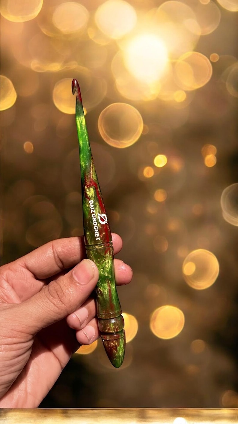 Puede incluir: Un ganchillo verde y rojo con el texto "DAIZ CROCHET" impreso en el lateral. El ganchillo se sostiene en una mano sobre un fondo borroso de luces bokeh doradas. El ganchillo mide aproximadamente 15 cm de largo.