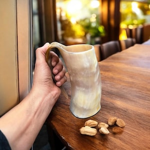Könnte beinhalten: Ein hornförmiger Becher mit gebogenem Griff und natürlicher, hellfarbiger Oberfläche. Der Becher wird von einer Hand gehalten. Mehrere Nüsse sind auf einem Holztisch verstreut. Der Becher ist etwa 15 cm hoch.