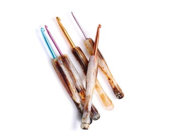 May include: A collection of crochet hooks with marbled brown and cream handles and metallic hook ends in various colors. The text "DAIZ CROCHET" is printed on the handles. The hooks are arranged on a white background.