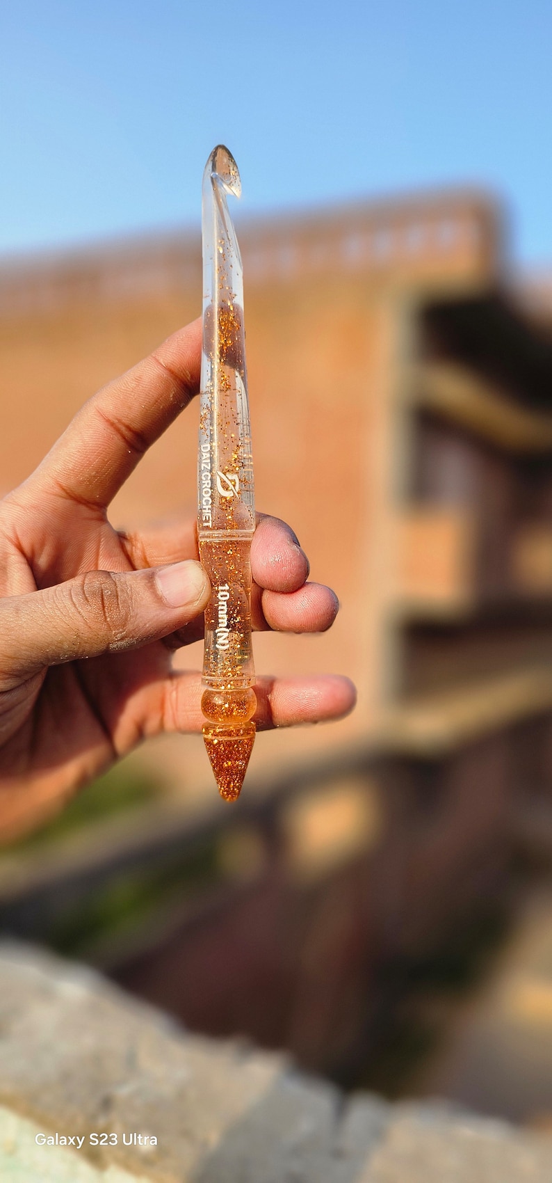May include: A clear resin crochet hook featuring gold glitter flakes embedded throughout the body. The tool displays the text DAIZ CROCHET and 10mm (N) printed in white on the shaft. The hook has a tapered point and a decorative pointed end.