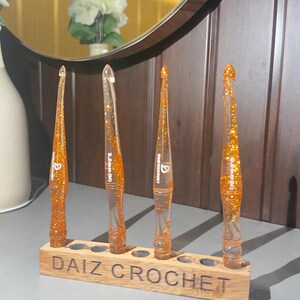 May include: Four orange resin crochet hooks with gold glitter embedded inside, each featuring a tapered handle and a hook tip. The hooks are displayed upright in a rectangular wooden stand with the text "DAIZ CROCHET" engraved on the front.