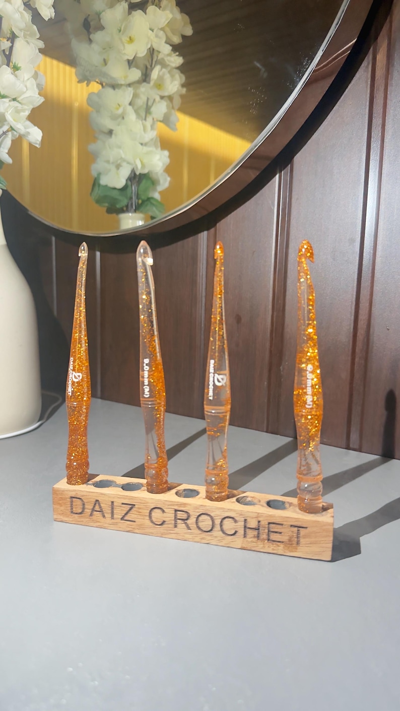 May include: Four orange resin crochet hooks with gold glitter embedded inside, each featuring a tapered handle and a hook tip. The hooks are displayed upright in a rectangular wooden stand with the text "DAIZ CROCHET" engraved on the front.