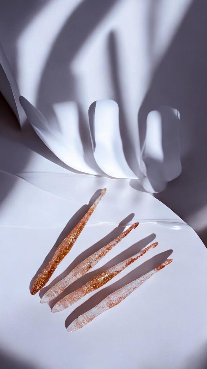 May include: A set of four translucent crochet hooks with embedded gold glitter particles. Each hook features a tapered head and a smooth, ergonomic handle. The text 5.5 mm is printed on the surface of one of the hooks, indicating its size.