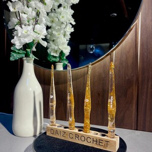 May include: Four clear resin crochet hooks filled with gold glitter, each featuring a specific size marking, standing upright in a rectangular wooden holder labeled DAIZ CROCHET. The hooks are arranged in a row on a black circular rotating display base.