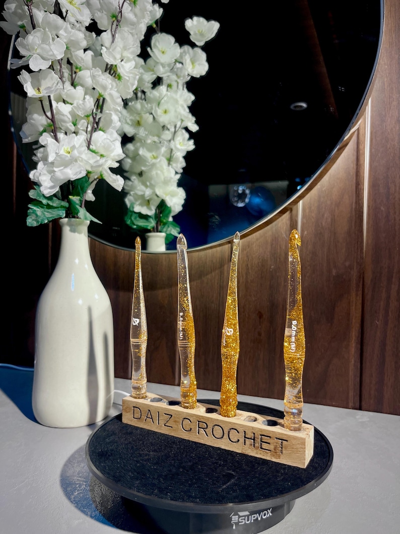 May include: Four clear resin crochet hooks filled with gold glitter, each featuring a specific size marking, standing upright in a rectangular wooden holder labeled DAIZ CROCHET. The hooks are arranged in a row on a black circular rotating display base.