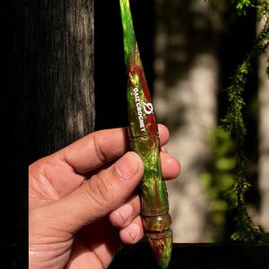 Puede incluir: Un ganchillo verde y rojo con el texto "DAIZ CROCHET" en blanco. El ganchillo tiene un efecto veteado y un mango redondeado. El ganchillo se sostiene en una mano.