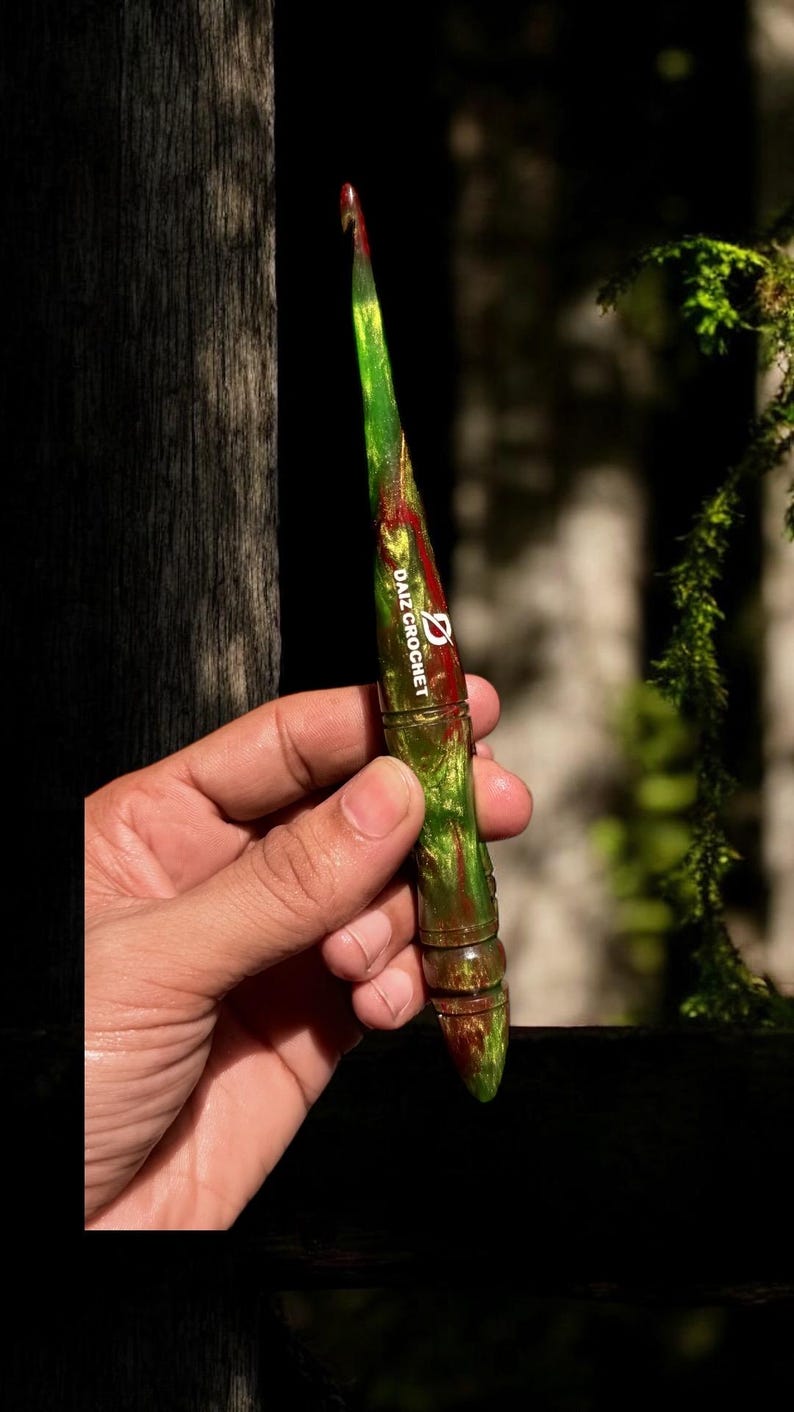 Puede incluir: Un ganchillo verde y rojo con el texto "DAIZ CROCHET" en blanco. El ganchillo tiene un efecto veteado y un mango redondeado. El ganchillo se sostiene en una mano.