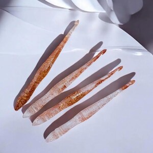 May include: Four translucent crochet hooks with embedded gold glitter particles. Each hook features a tapered head and a textured grip area. The hooks are arranged diagonally, with the size 6.5mm clearly printed on the surface of the third hook.