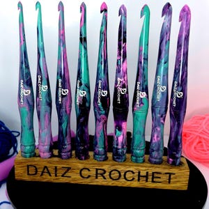 Puede incluir: Un juego de nueve ganchillos de crochet coloridos con un dise&ntilde;o de m&aacute;rmol en tonos de rosa, verde y negro. Los ganchillos se muestran en un soporte de madera con el texto "DAIZ CROCHET" grabado en la parte delantera.