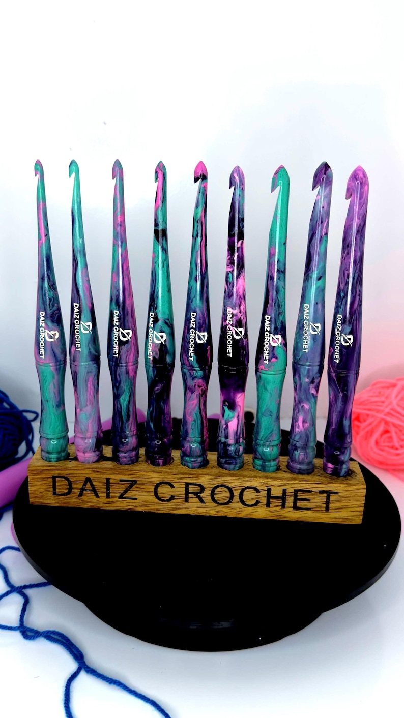 Puede incluir: Un juego de nueve ganchillos de crochet coloridos con un dise&ntilde;o de m&aacute;rmol en tonos de rosa, verde y negro. Los ganchillos se muestran en un soporte de madera con el texto "DAIZ CROCHET" grabado en la parte delantera.