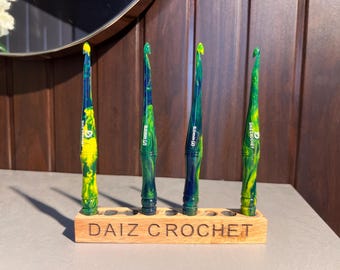 May include: Four crochet hooks with marbled yellow, green, and blue resin handles standing in a rectangular wooden stand. The stand is engraved with the text DAIZ CROCHET. Two hooks are labeled 6.0mm (J) and two are labeled DAIZ CROCHET.