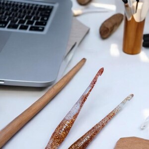 May include: Two clear resin crochet hooks with embedded copper-colored glitter, each featuring the text 5.5mm printed on the shaft. The hooks have a tapered design and are positioned diagonally on a white surface near a laptop and wooden tools.