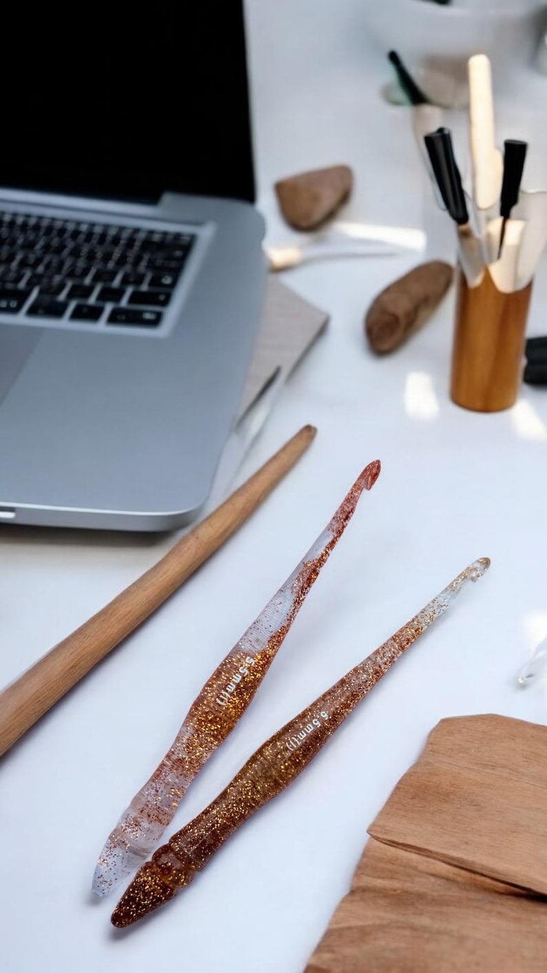 May include: Two clear resin crochet hooks with embedded copper-colored glitter, each featuring the text 5.5mm printed on the shaft. The hooks have a tapered design and are positioned diagonally on a white surface near a laptop and wooden tools.