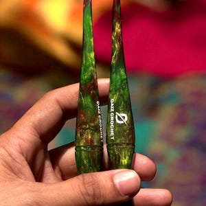 Puede incluir: Dos ganchillos verdes y marrones con el texto "DAIZ CROCHET" impreso. Los ganchillos tienen un efecto veteado y se sostienen en una mano. Los ganchillos son para tejer.