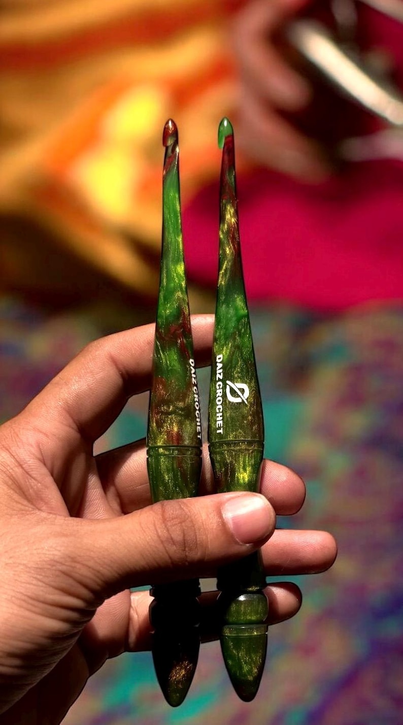 Puede incluir: Dos ganchillos verdes y marrones con el texto "DAIZ CROCHET" impreso. Los ganchillos tienen un efecto veteado y se sostienen en una mano. Los ganchillos son para tejer.