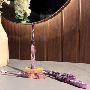 Puede incluir: Un conjunto de cuatro ganchillos con un acabado de resina veteada en color morado y blanco. Un ganchillo se mantiene vertical en un pequeño bloque de madera, mientras que otros tres yacen horizontalmente. Cada uno muestra el nombre Tazimont.