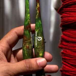 Puede incluir: Dos ganchillos con vetas verdes, rojas y doradas. Los ganchillos tienen un dise&ntilde;o c&oacute;nico y el texto "DAIZ CROCHET" est&aacute; impreso en el mango. Los ganchillos se sostienen en una mano.