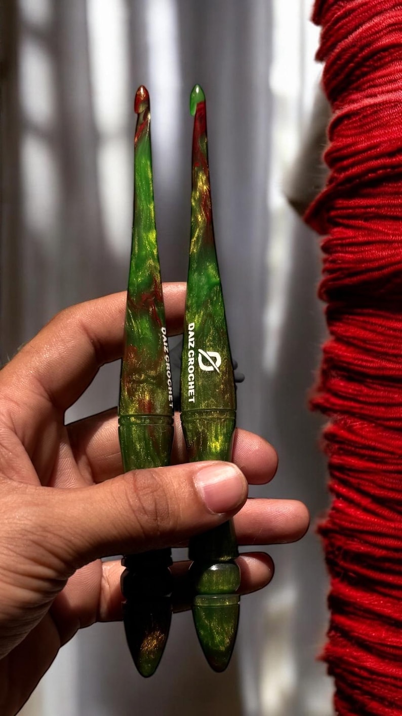 Puede incluir: Dos ganchillos con vetas verdes, rojas y doradas. Los ganchillos tienen un dise&ntilde;o c&oacute;nico y el texto "DAIZ CROCHET" est&aacute; impreso en el mango. Los ganchillos se sostienen en una mano.