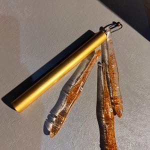 May include: A set of three clear resin crochet hooks filled with gold glitter, each featuring the text DAIZ CROCHET, accompanied by a cylindrical gold metal storage case with a black cord and a small wooden bead attached to the end.