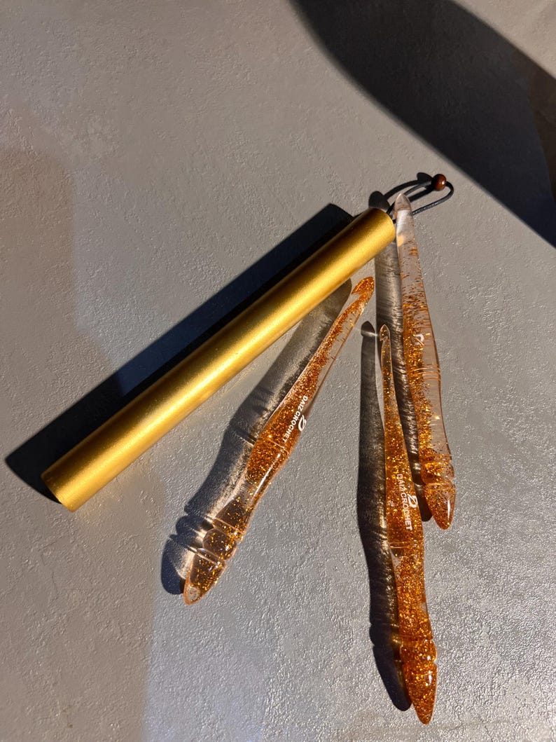 May include: A set of three clear resin crochet hooks filled with gold glitter, each featuring the text DAIZ CROCHET, accompanied by a cylindrical gold metal storage case with a black cord and a small wooden bead attached to the end.
