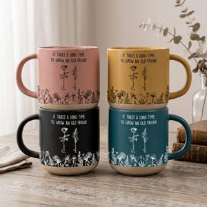 Pode incluir: Quatro canecas de cerâmica empilhadas, cada uma de uma cor diferente: rosa, amarelo, preto e verde-azulado. Cada caneca apresenta o texto "IT TAKES A LONG TIME TO GROW AN OLD FRIEND" com um design floral. As canecas têm uma base bege e uma alça curva.