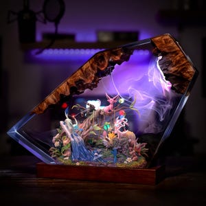 May include: A decorative resin sculpture featuring a fantasy scene with colorful figures, set within a clear, angled frame. The frame has a wooden top and base. The scene is illuminated with purple and white lights, creating a magical ambiance.