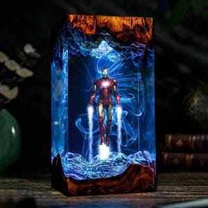May include: A rectangular resin art piece featuring an Iron Man figure. The figure is red and gold, with blue energy effects. The resin block has a wooden base and top, with blue and white swirling patterns throughout.