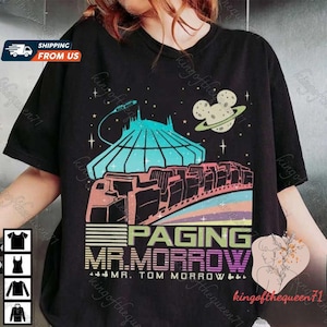 Könnte beinhalten: Schwarzes T-Shirt mit einer bunten Grafik eines futuristischen Zuges, der durch den Weltraum fährt. Die Grafik enthält eine Raumstation, einen Planeten mit Mickey-Maus-Ohren und den Text "PAGING MR. MORROW MR. TOM MORROW &..."