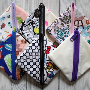 May include: A collection of small, square fabric pouches with zippers in various colors. The pouches feature diverse patterns, including floral, geometric, and cartoon designs. The zippers are in contrasting colors like purple, blue, and black.