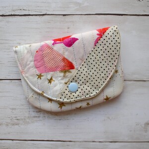 May include: A quilted fabric pouch featuring a white base with gold star patterns and a flap decorated with pink fruit illustrations. The flap has a black polka dot section and is secured with a single light blue plastic snap button closure.