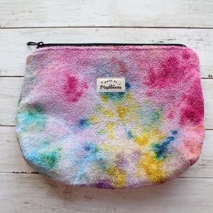 May include: A zippered pouch made of a textured, terry cloth-like fabric featuring a vibrant tie-dye pattern in shades of pink, yellow, blue, and purple. A small rectangular white label is centered on the front with the text Made by Maraboots.