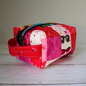 May include: A patchwork fabric cosmetic bag in shades of red, pink, and white. The bag features a black zipper and a red handle. One side has a whimsical design of a face with sunglasses and a bow tie. The bag is approximately 7 inches long.