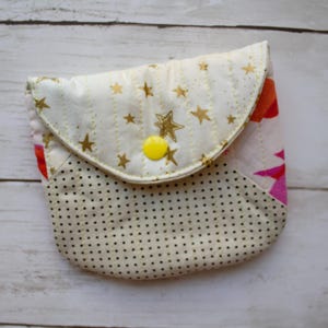 May include: A quilted fabric pouch featuring a cream-colored flap with gold foil stars and a bright yellow snap button. The lower section of the pouch is cream with a black polka dot pattern, and a glimpse of pink and orange floral fabric is visible.