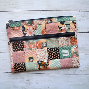 May include: A rectangular fabric zipper pouch featuring a patchwork design with various patterns including pumpkins, black cats, flowers, stars, and candy corn in shades of orange, green, black, and cream. It has two black zippers and a small white label.