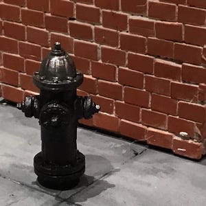 May include: A black fire hydrant with a silver top stands on a gray surface in front of a red brick wall. The hydrant has multiple nozzles and a round base. The brick wall is made of rectangular bricks.