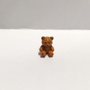 May include: A small, brown teddy bear figurine. The bear is sitting upright with its arms and legs slightly spread. The bear's fur is a warm brown color, and it has small, round ears and a detailed face. The figurine is on a white surface.