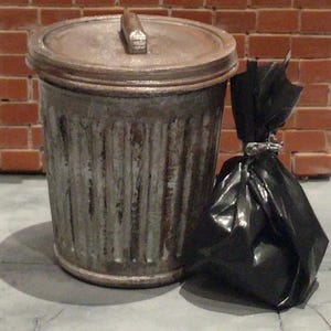 May include: A weathered metal trash can with a lid and handle, next to a tied-up black trash bag. The can has vertical ridges and a rusty appearance. The background features a brick wall and a concrete surface.