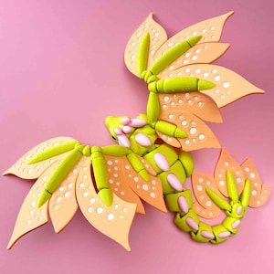 Op de afbeelding: Een 3D-geprint drakenbeeldje met een groen lichaam en roze en oranje vleugels. De draak heeft een gesegmenteerd lichaam en staat in een vliegende houding.