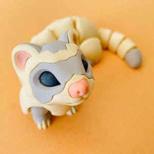 Puede incluir: Una pequeña figurita de hurón segmentada, blanca y gris, con la nariz rosa y los ojos negros. La figurita está en posición enroscada, con la cola envuelta alrededor del cuerpo.