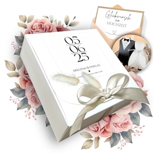 May include: A white rectangular gift box featuring the date 05.06.25, the names Melina & Niklas, and the text Hand in Hand ein Leben lang. The box is decorated with a beige ribbon, dried grass, and a card reading Gluckwunsch zur Hochzeit.