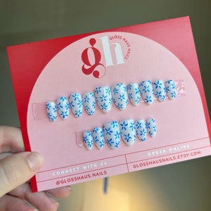 May include: A set of 10 white press-on nails with a blue floral design. The nails are displayed on a pink and red card with the text "Gloss Haus Nails" and "Connect with us @glosshaus.nails".