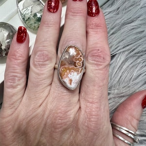 May include: A silver ring with a large, oval-shaped stone. The stone is a mix of white, brown, and orange colors with a swirling pattern. The ring is on a person's finger.