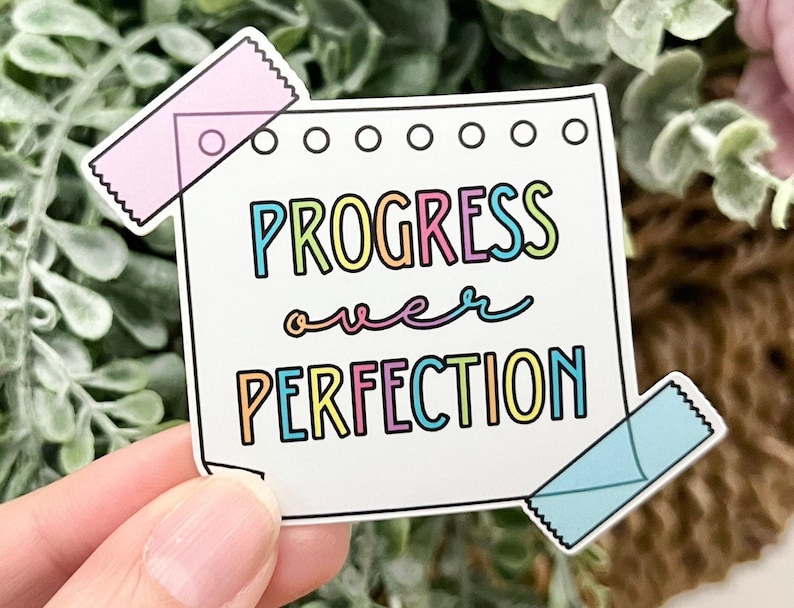 May include: A white sticker with a colorful design that says "Progress over Perfection" with a pink and blue washi tape on the top and bottom.