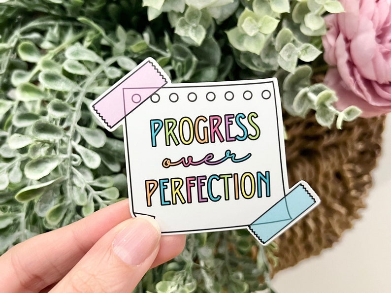 May include: A white sticker with the text "PROGRESS over PERFECTION" in rainbow colors. The sticker has a pink and blue washi tape on each side.