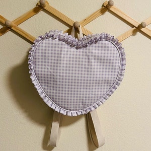 May include: A heart-shaped purple and white gingham backpack with a ruffled trim and adjustable straps.