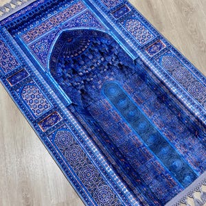 Op de afbeelding: Een blauw gebedskleed met een ingewikkeld, sierlijk ontwerp. Het kleed heeft een centraal boogmotief en gedetailleerde geometrische patronen in blauw- en wittinten. De randen zijn afgewerkt met kwastjes.