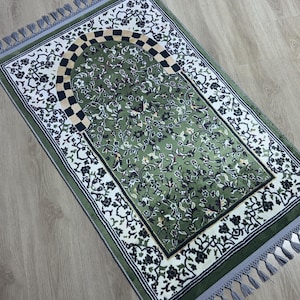 May include: A green prayer rug with a floral design and a decorative arch. The rug has a white border with black floral accents and gray tassels on the edges. The central field is a darker green with a repeating floral pattern.