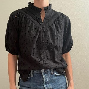 May include: Black eyelet blouse with a high ruffled collar and short, puffed sleeves. The blouse has a button-down front and is paired with blue denim jeans. The top features a delicate, openwork design.