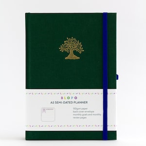 Puede incluir: Un planificador A5 semidateado de color verde con un diseño de árbol dorado en la portada. El planificador tiene una banda elástica azul e incluye papel de 150 g/m², un sobre de contraportada, objetivos mensuales y páginas de revisión mensuales.