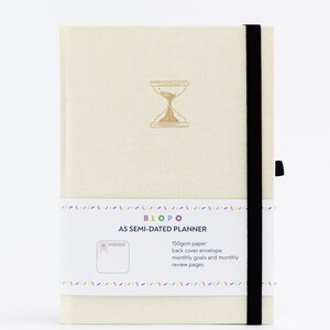 Puede incluir: Una agenda A5 semidiaria de color crema con cierre elástico negro. La portada presenta un gráfico de reloj de arena dorado y el texto "BLOPO A5 SEMI-DATED PLANNER". La parte posterior de la agenda tiene un bolsillo e incluye papel de 150 g/m², objetivos mensuales y páginas de revisión mensuales.